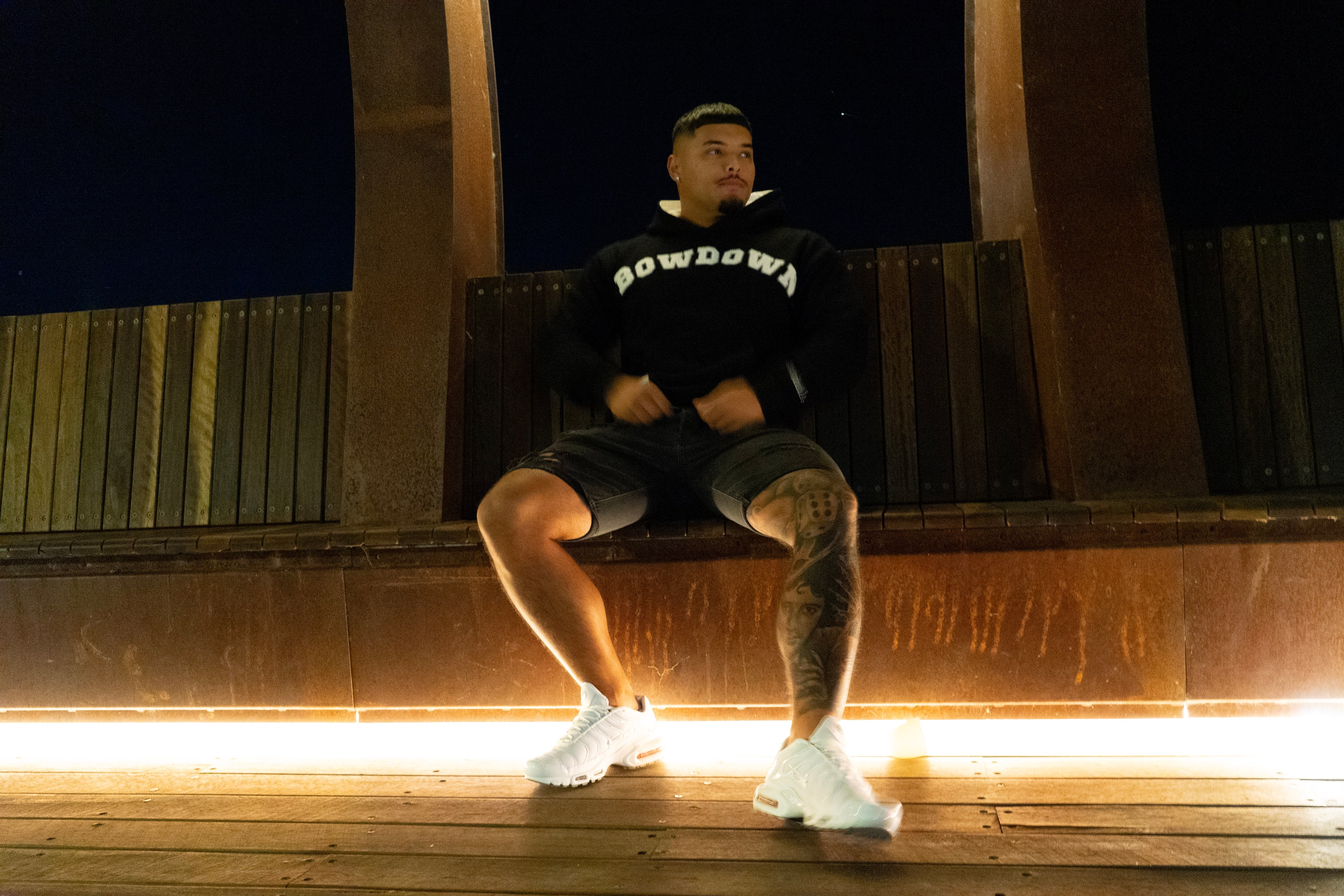 Man sitting on a wooden bench at night wearing a black sweatshirt with 'BOWDOW' text.